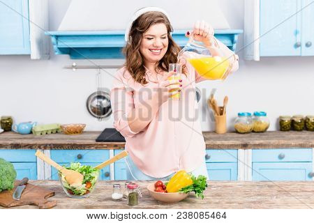 Smiling Overweight Woman In Headphones Pouring Juice Into Glass In Kitchen