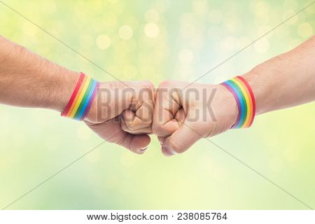 lgbt, same-sex love and homosexual relationships concept - close up of male couple hands with gay pride rainbow awareness wristbands making fist bump gesture over green lights background