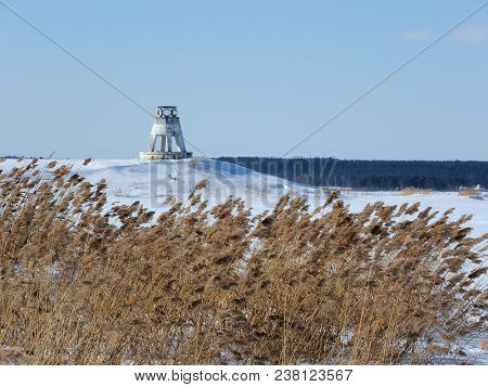 Winter Landscape. A Small Lighthouse On The Bank Of A Frozen River. Reeds In The Foreground.