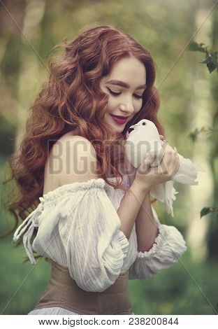 A Sensual Red-haired Girl In A White Dress With Bare Shoulders Holds A White Dove In Her Hand. White