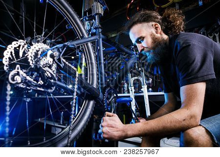 Young Stylish Bearded, With Long Hair Caucasian Male Mechanic Worker At A Bicycle Workshop Uses A To