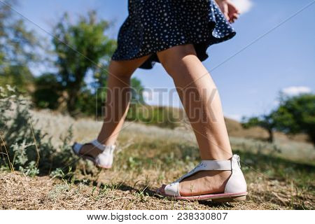 A Girl In Sandals And A Blue Dress Is Walking Along The Sun-lit Grass