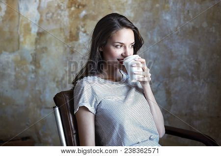 Charming Young Brunette Lady Relaxing Indoors, Drinking Coffee, Having Joyful Look. Pretty Girl With