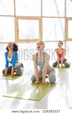 Group of active girls sitting in pose of lion, rolling eyes and sticking out tongues during yoga practice in gym