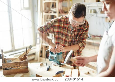 One of female carpenters working with electric fretsaw by workbench while her co-worker hammering nails