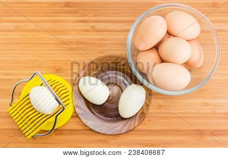 Top View Of Egg Slicer With Egg In It, Two Boiled Peeled Eggs On Saucer And Not Cleaned Eggs In A Gl