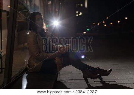 Girl Brunette Sitting Near In The Shopwindow On The Night City Street In A Cold Time