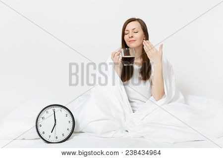 Happy Woman Sitting In Bed With Round Clock, White Sheet, Pillow, Wrapping In Blanket On White Backg