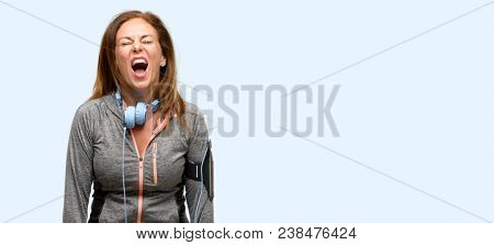 Middle age gym fit woman with workout headphones stressful, terrified in panic, shouting exasperated and frustrated. Unpleasant gesture. Annoying work drives me crazy isolated blue background