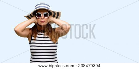 Middle age cool woman wearing summer hat and sunglasses covering ears ignoring annoying loud noise, plugs ears to avoid hearing sound. Noisy music is a problem. isolated blue background