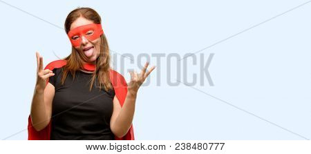 Middle age super hero woman wearing red mask and cape looking at camera showing tong and making victory sign with fingers isolated blue background