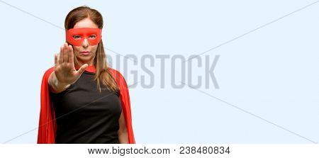 Middle age super hero woman wearing red mask and cape annoyed, bad attitude making stop sign with hand, saying no, expressing security, defense or restriction, maybe pushing isolated blue background