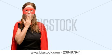 Middle age super hero woman wearing red mask and cape covers mouth in shock, looks shy, expressing silence and mistake concepts, scared isolated blue background