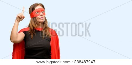 Middle age super hero woman wearing red mask and cape raising finger, the number one isolated blue background
