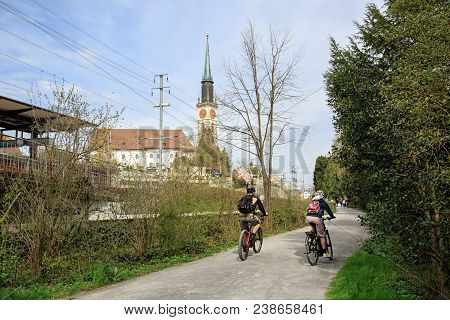 Cham, Switzerland - April 15, 2018. A Young Couple Riding Bicycles In The Villette-park In Front Of 