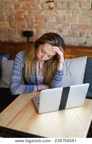 Young Woman Sitting In Cozy Coffee House Near Wooden Table With Glass Cup Of Cappuccino And Using La