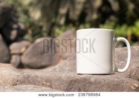 White Blank Coffee Mug Mock Up, Close-up Of Mug Outside In The Sunshine On Some Large Decorative Gar