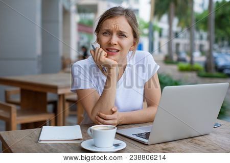 Portrait Of Frowning Young Girl Sitting With Laptop At Cafe. Displeased Young Caucasian Freelancer O