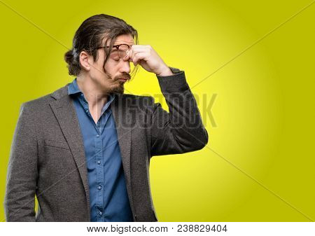 Handsome young man with sleepy expression, being overworked and tired, rubbes nose because of weariness