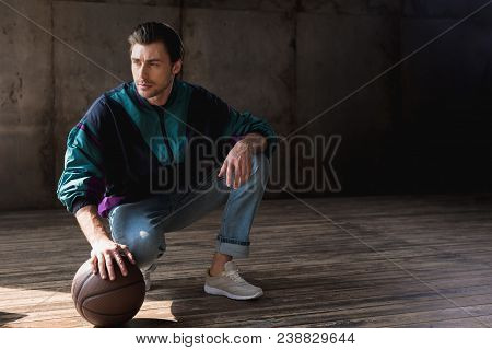Handsome Young Man In Vintage Windcheater With Basketball Ball Sitting Squats On Wooden Floor