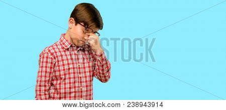 Handsome toddler child with green eyes with sleepy expression, being overworked and tired, rubbes nose because of weariness over blue background