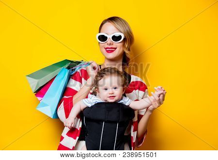 Mother And Little Baby In Carrier Have A Shopping With Bags. Models On Yellow Background