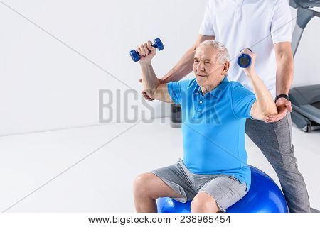 Partial View Of Rehabilitation Therapist Assisting Senior Man Exercising With Dumbbells On Fitness B