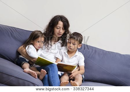 Family Ideas And Concepts. Mother Reading A Book Together With Her Little Children On Couch Indoors.
