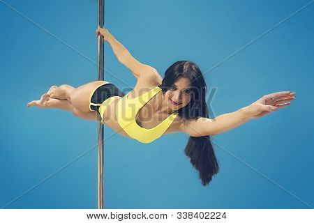 Young Sexy Brunette Making Superman Element Hanging On Pole At Blue Background. Fit Smiling Poledanc