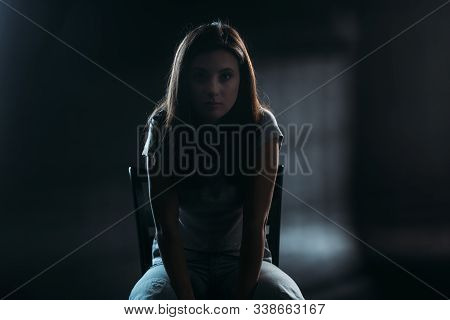 Hopeless Young Woman Looking At Camera While Sitting On Chair In Darkness On Black Background
