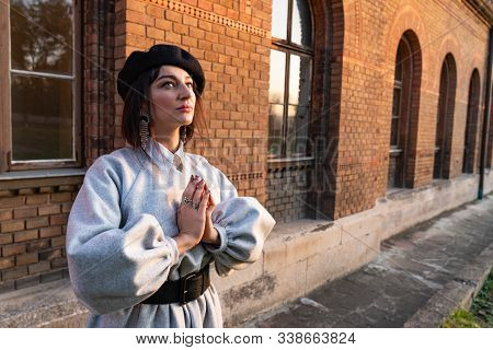 Calmy Brunette Girl In Grey Dress And Black Cap Holds Her Hands Together And Looking Up, Red Brick W