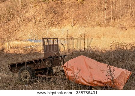 Old Rusted Two Wheel Farm Trailer In Field Of Tall Brown Grass Next To An Orange Canvas Bag.