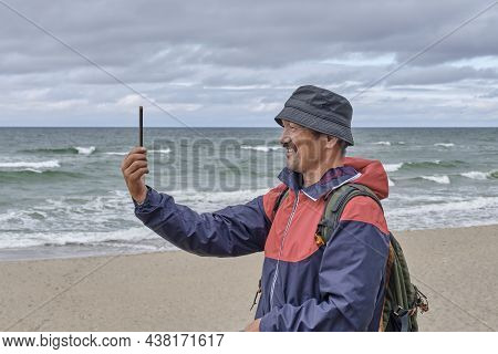 Senior Asian Man In Panama Hat, In Red And Blue Windbreaker, With Backpack Standing On Seashore, Usi