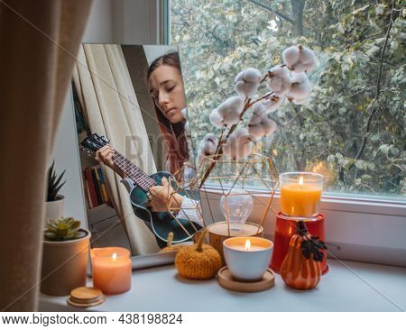 Girl Sits In Her Room On A Window Sill Decorated For The Holiday And Plays The Ukulele. Her Reflecti