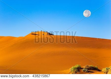 The huge moon is visible low above the horizon. Huge scenic red and orange sand dunes. Namibia.  Sossusflei is a clay plateau in the Namib Naukluft Park. Africa. 