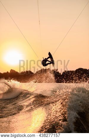 Beautiful Dark Silhouette Of Man Holding Rope And Making Jump On Wakeboard At Sunset.