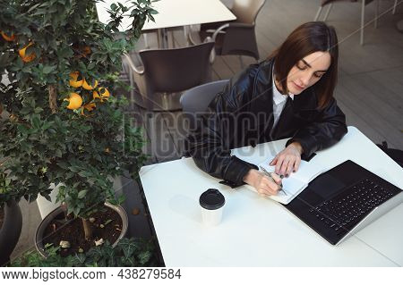 Overhead View Of Concentrated Woman, Copywriter Writing On Notepad, Using Laptop Sitting At Table Wi