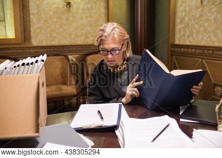 Caucasian female lawyer with big open dictionary of juridical terms or encyclopaedia looking though financial document by workplace