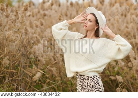 Elegant young woman in a white woolen sweater and a white felt hat poses in the autumn park on the background of reeds. Autumn fashion and beauty. 