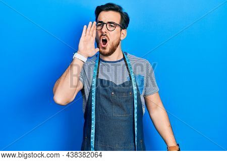 Handsome caucasian man with beard dressmaker designer wearing atelier apron shouting and screaming loud to side with hand on mouth. communication concept. 