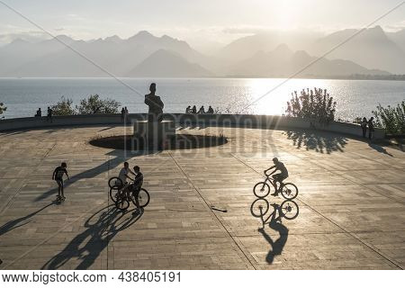 Antalya, Turkey - October 10, 2020: Unidentified kids cycling bicycles in front of a sculpture monument of a worker and son in Karaalioglu Park at sunset over mountains and the sea in Antalya, Turkey
