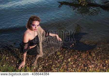 Passionate Mermaid Posing With Net Near Sea On Rocks While Wearing Artistic Makeup And Facial Strass