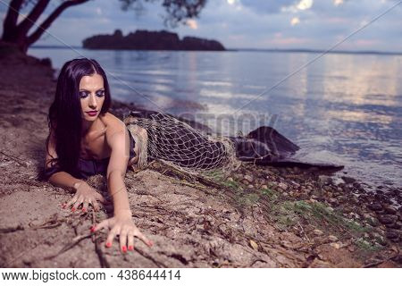 Mermaid Posing With Net Near Sea On Rocks While Wearing Artistic Makeup And Black Shiny Tail Covered