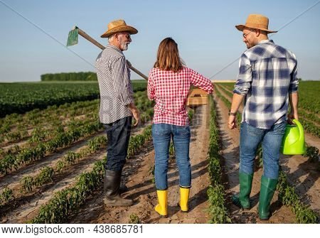 Elderly Farmer Wearing Straw Hat Standing In Field With Gardening Hoe On Shoulder. Young Woman Holdi