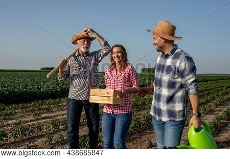 Elderly Farmer With Gardening Hoe On Shoulder Holding Straw Hat With Hand. Young Woman Carrying Wood