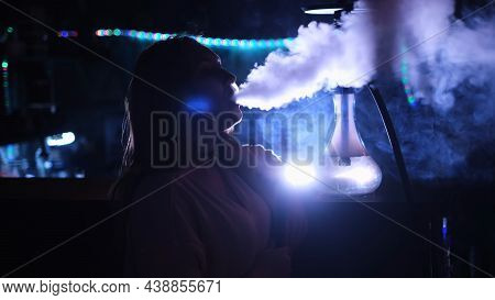 Close Up Side View Of A Girl Smoking Hookah On Blue Neon Background. Media. Female While Smoking A H