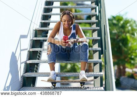 Black Woman With Coloured Braids, Consulting Her Smartphone With Her Feet Resting On A Skateboard.