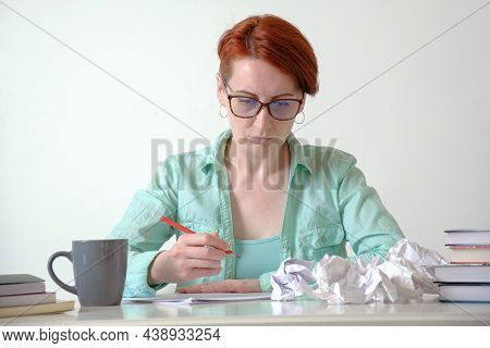 A Young Woman At A Desk Makes Notes On Paper With A Red Pencil.