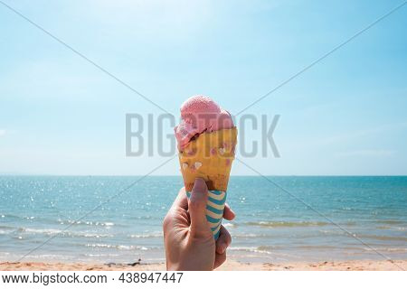 Strawberry Ice Cream In A Wafer Cone Sticking Out Held In Woman`s Hand With Tropical Beach Backgroun