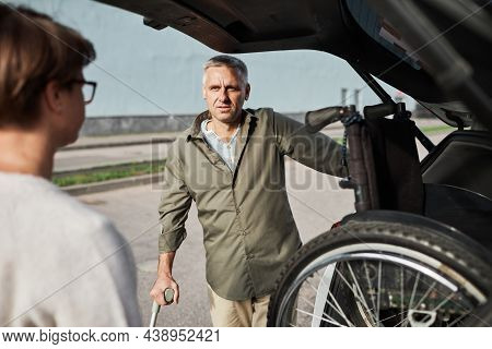 Portrait Of Mature Couple Unloading Wheelchair Out Of Car Trunk In Parking Lot Outdoors, Copy Space